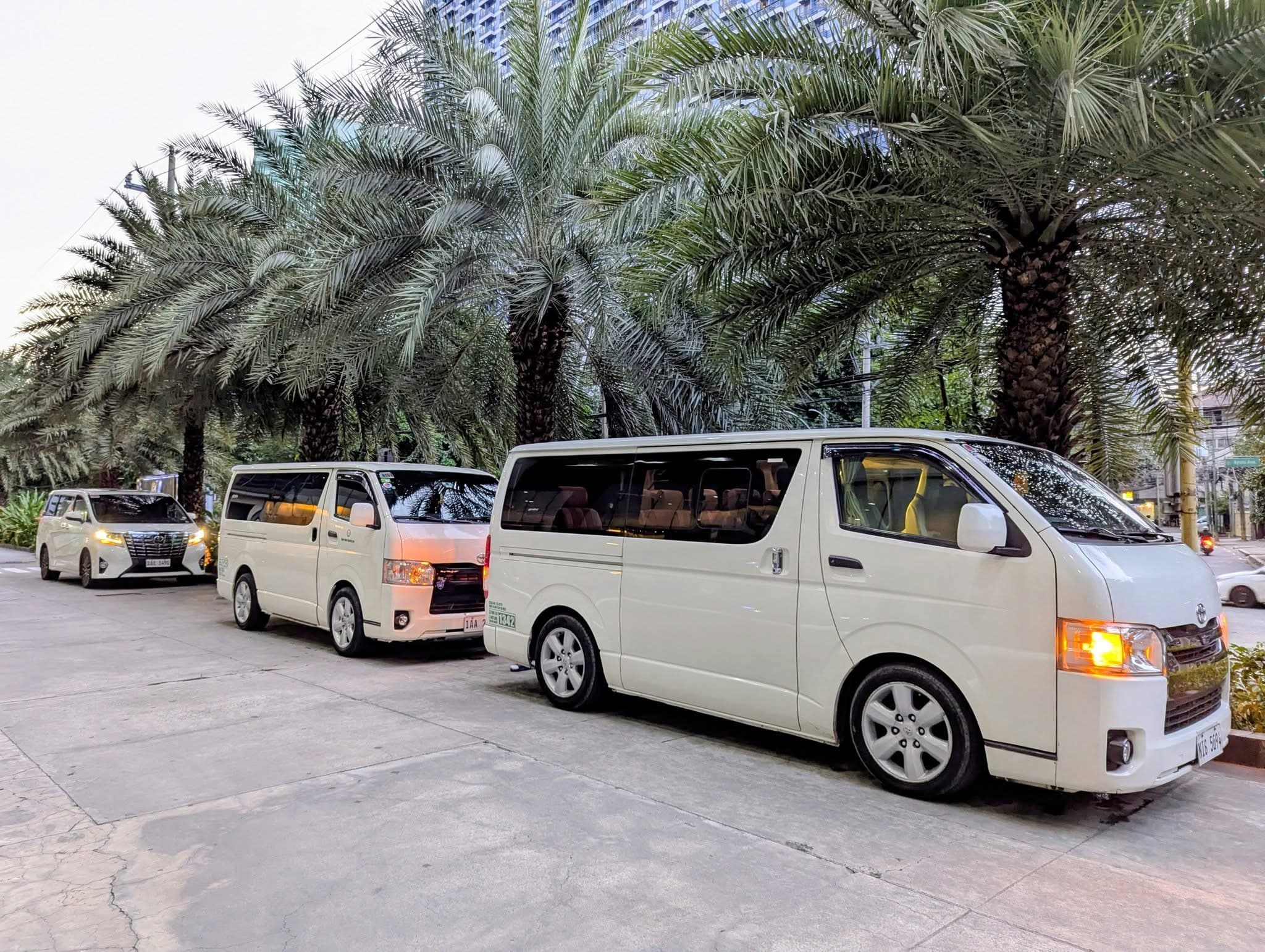 Three white Toyota Hiace vans parked in a row next to lush green palm trees.