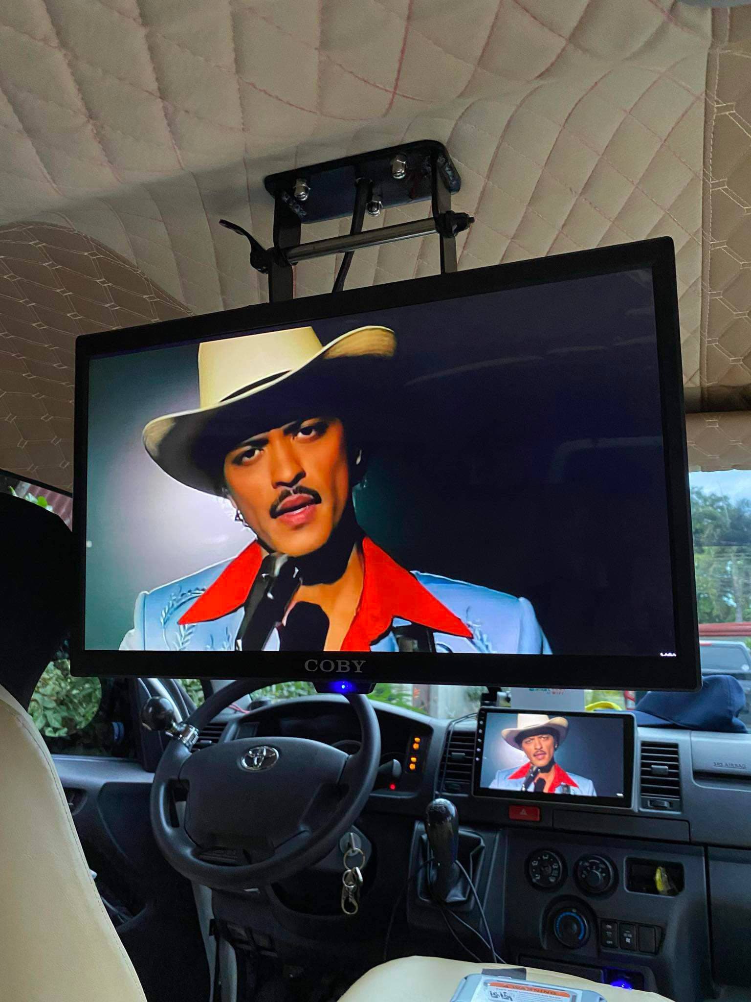 Ceiling-mounted monitor and dashboard screen in a vehicle showing a man in a cowboy hat.