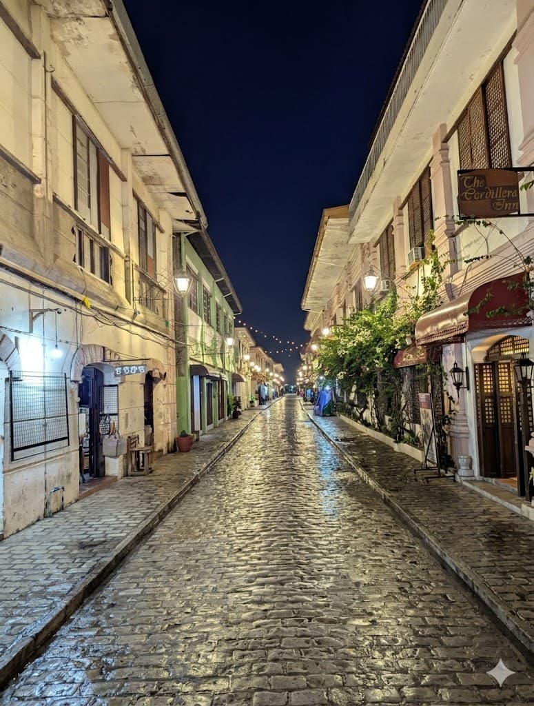 Narrow wet cobblestone street at night lined with historic colonial buildings and glowing lamps.