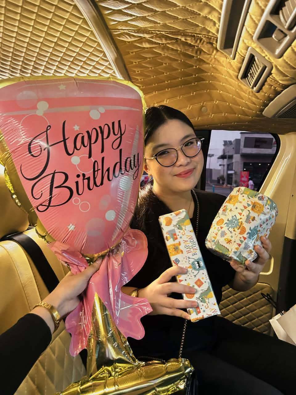 Smiling woman with glasses holding birthday gifts and a pink balloon inside a gold-trimmed vehicle.