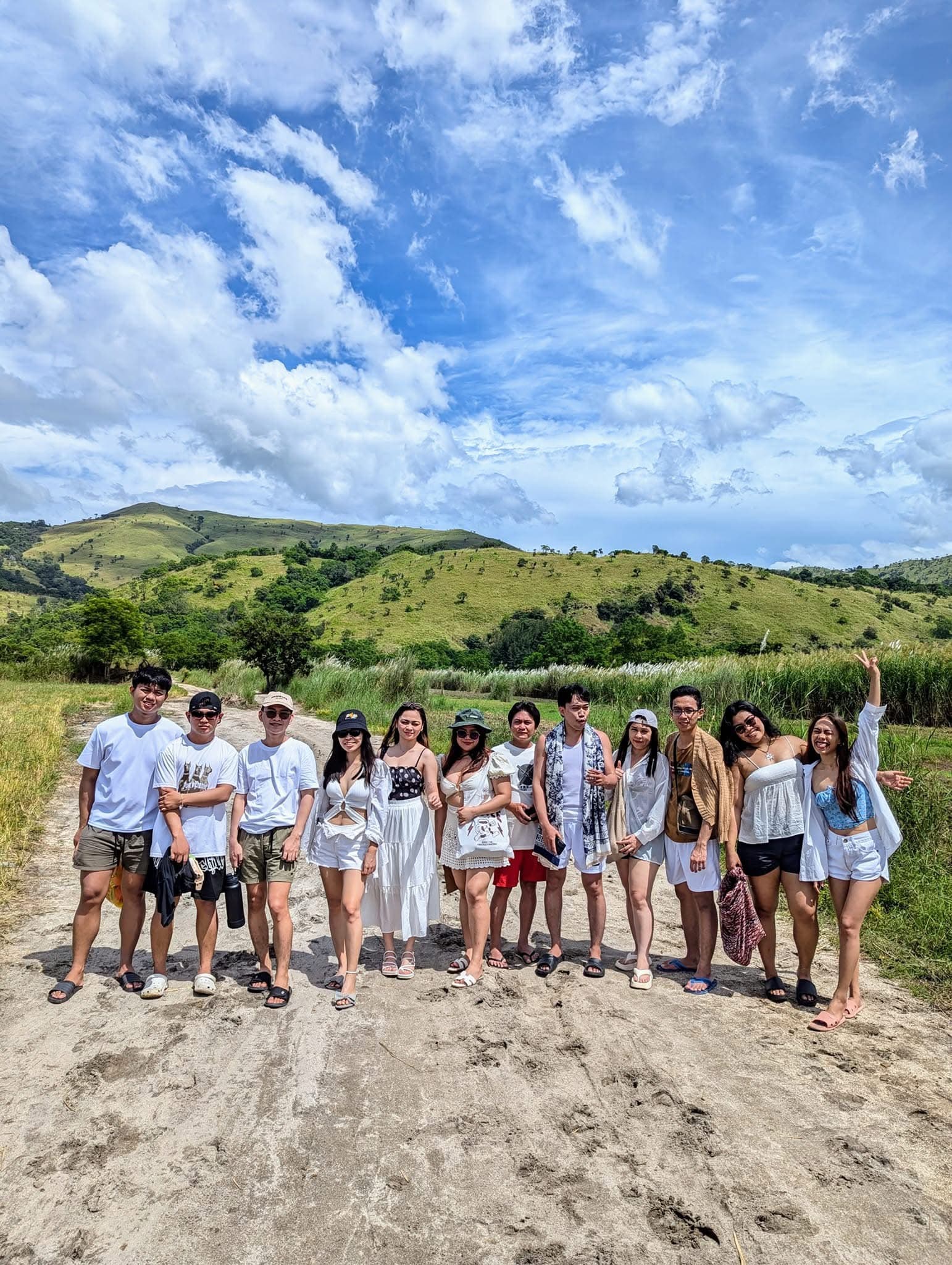 Friends posing on a dirt path with scenic green hills and a bright cloudy sky.