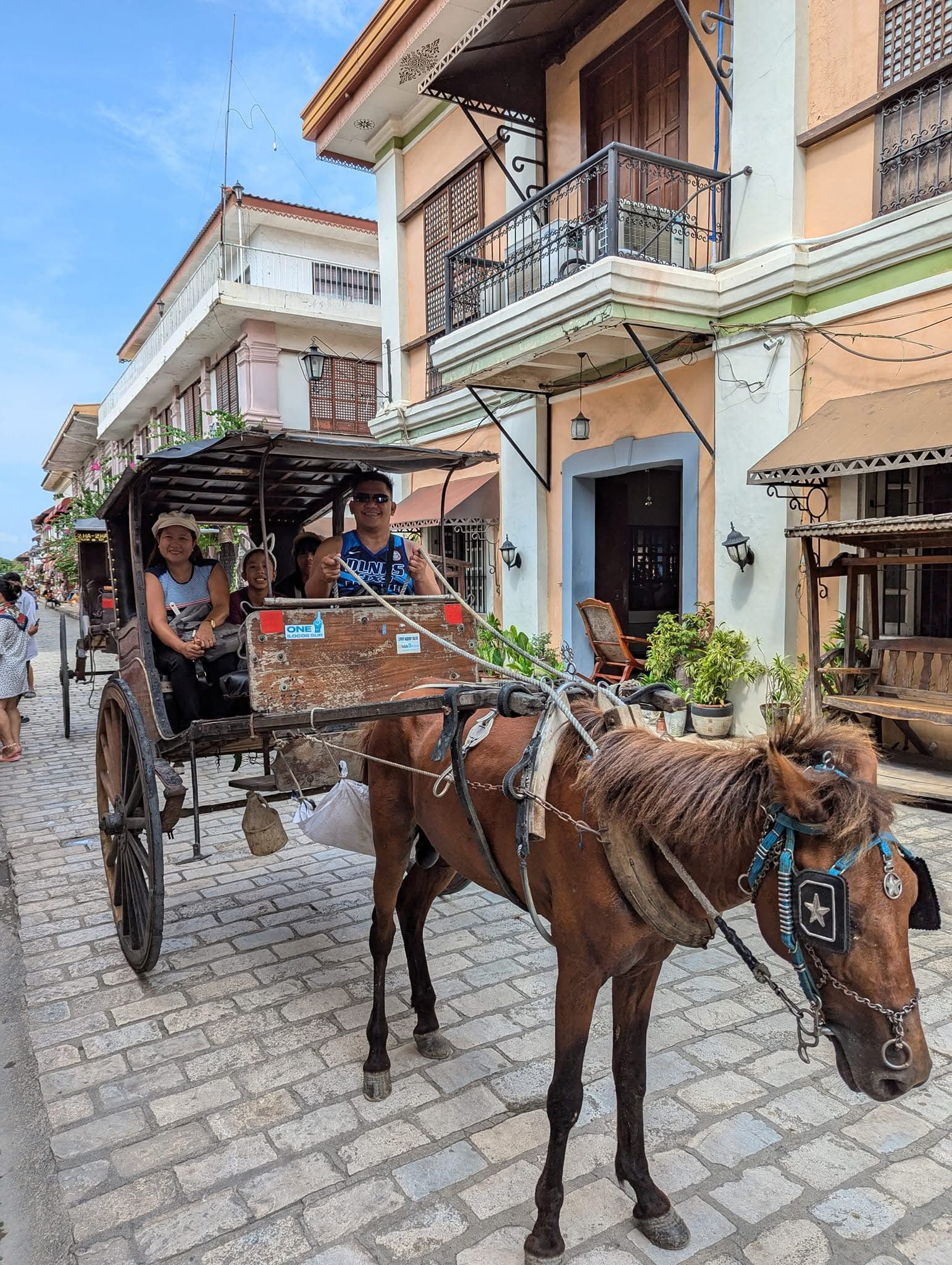 A family rides a horse-drawn carriage along a historic cobblestone street with colonial buildings.
