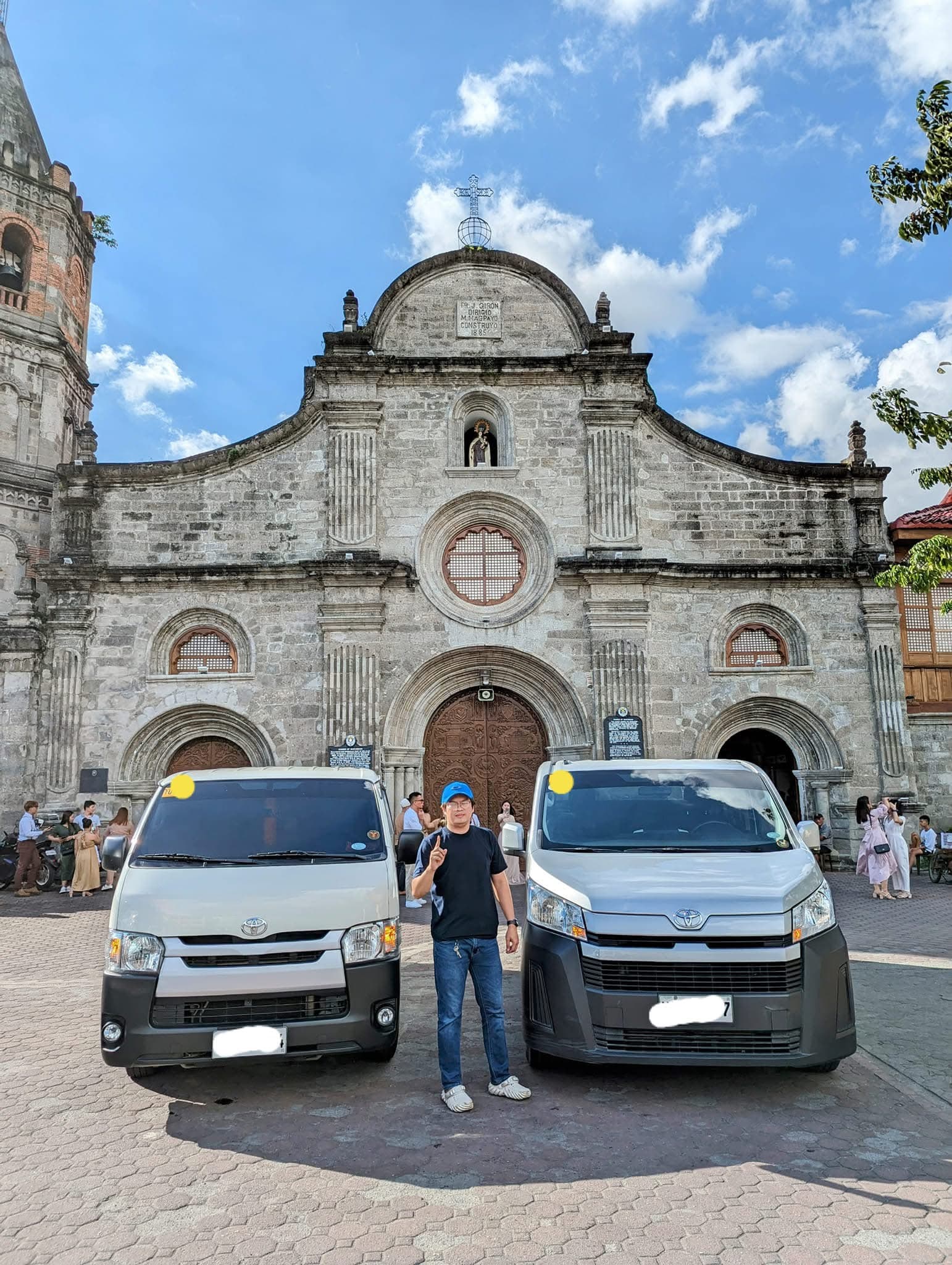 Man in blue cap stands between two white vans before a historic stone church.